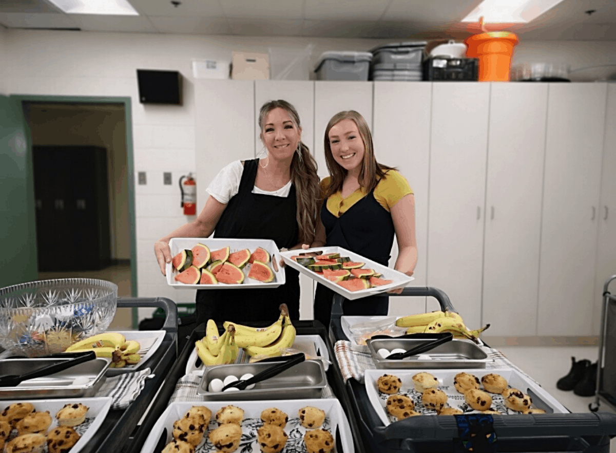 Two women ready to serve breakfast with trays full of eggs, bananas and muffins